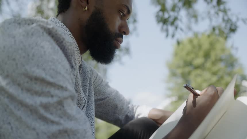 A man sits under a tree and writes in a notebook