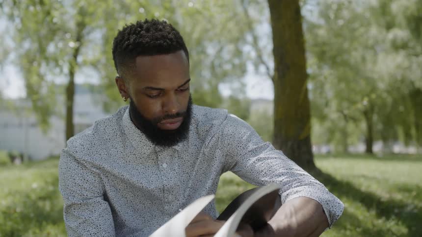 A bearded man sits on a lawn and writes in his notebook