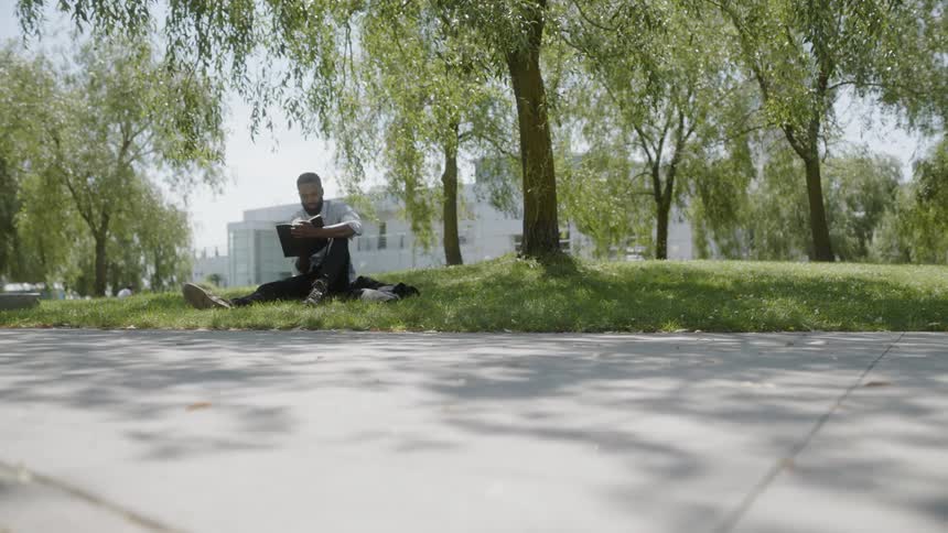 A man reads a book while sitting on the grass beneath a large shade tree