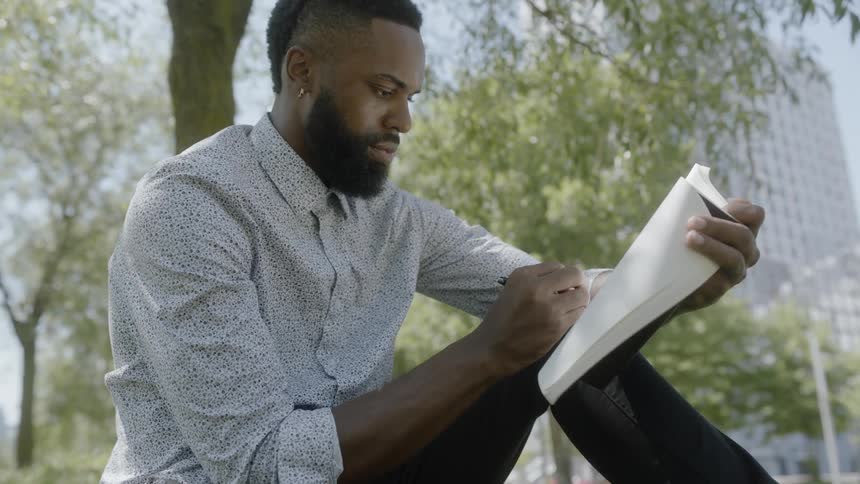 A young black man sits outside and writes in a book