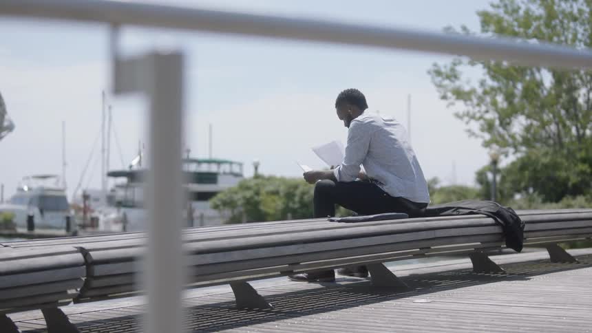 Black man sitting on bench overlooking the marina reading documents