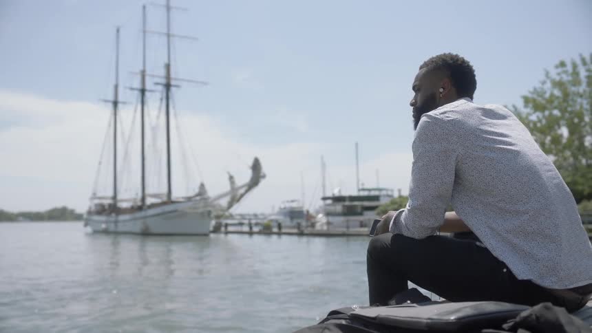 A black man sitting on a bench at the docks listening to earphones