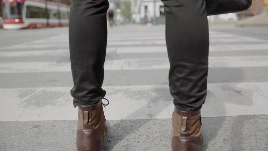 A man dressed in skinny black pants and brown leather boots stand at the crosswalk waiting to cross