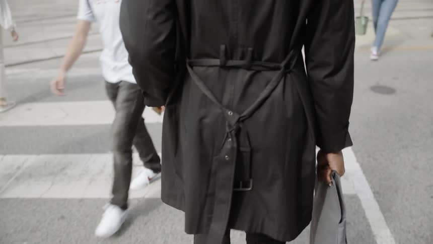 A young fashionably dressed man walks across a city street carrying a portfolio