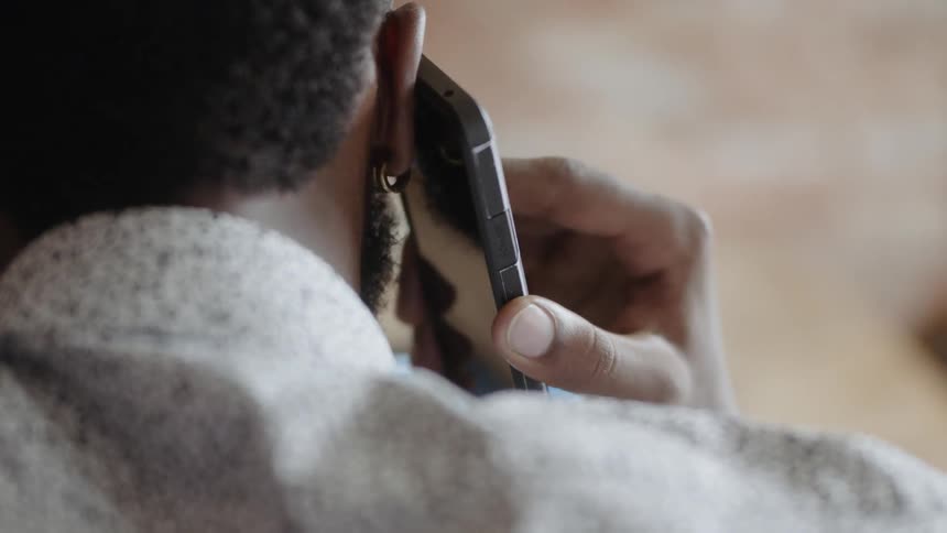 Black man wearing earrings uses laptop while talking on a cell phone by day