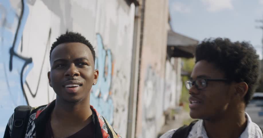 Black man who wears glasses talks to another black man who has earrings on while walking in the city by day