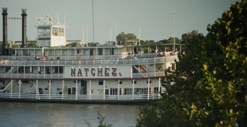 Riverboat paddles down river past trees and buildings