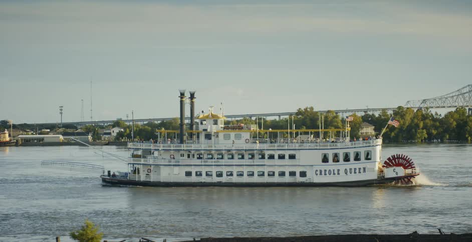 A white steam powered paddle boat cruises along a large river as passengers are seen walking along the upper deck on a clear day