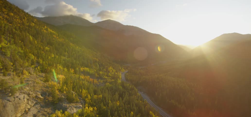 The sun shines brightly over a forested mountain range with a road cutting through it