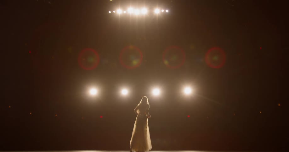 A woman in a long dress performs on a stage underneath stage lights
