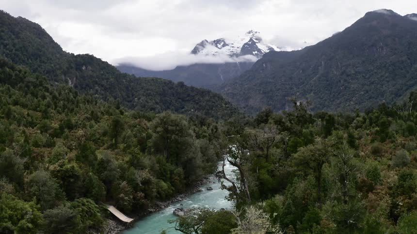 A mountain stream running through a forest with snow covered peaks and clouds above