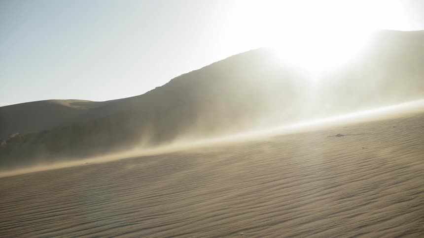 Sand blows around dunes with a bright sun behind