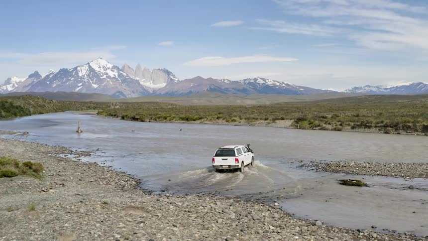 Car drives through the creek with the landscape of snow capped mountains by day