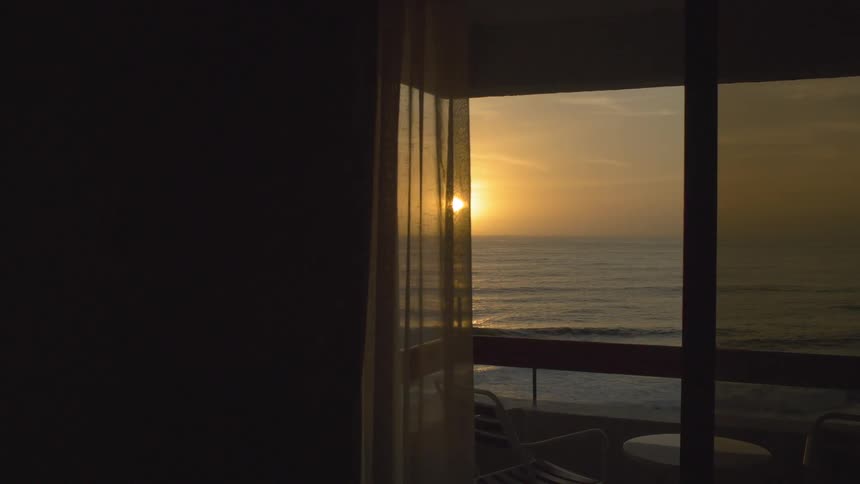 The camera looks out an open door to the sun setting over the ocean