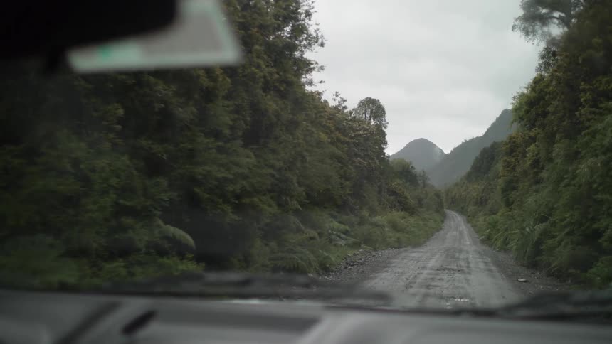 A vehicle's windshield wipers go back and forth as it travels down a narrow gravel road densely lined with trees