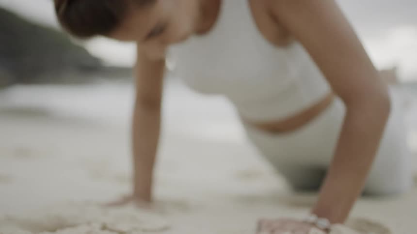 A woman in a white sports bra and yoga pants does push ups on a sandy beach