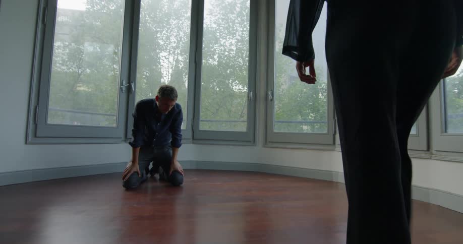 A man who is sitting on a hardwood floor in front of a bay window gets up on his knees while a woman approaches him and stops to stare down at him intensely