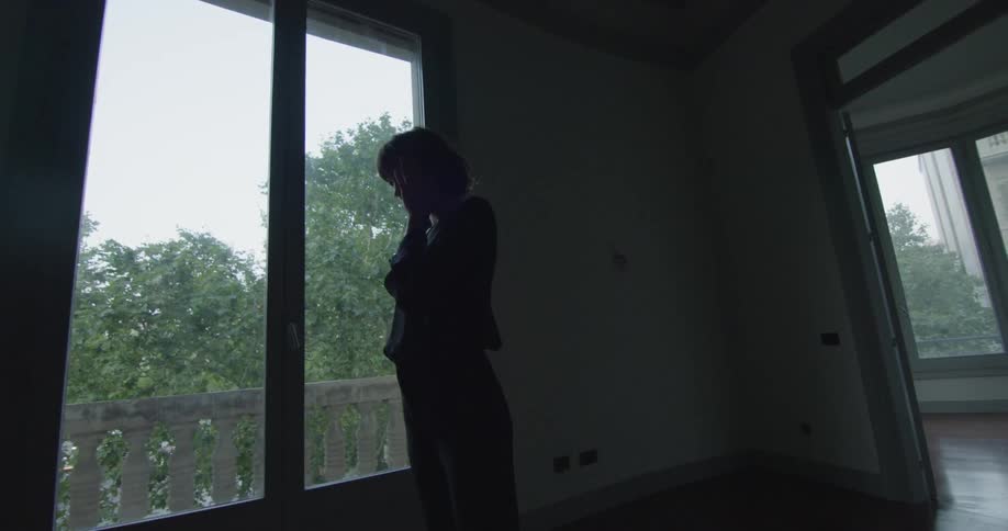 A woman wiping her eyes while standing in front of a window in a dimly lit room