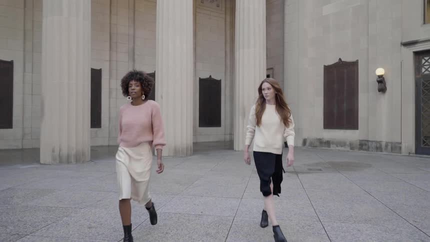 Two women strut forward away from building columns