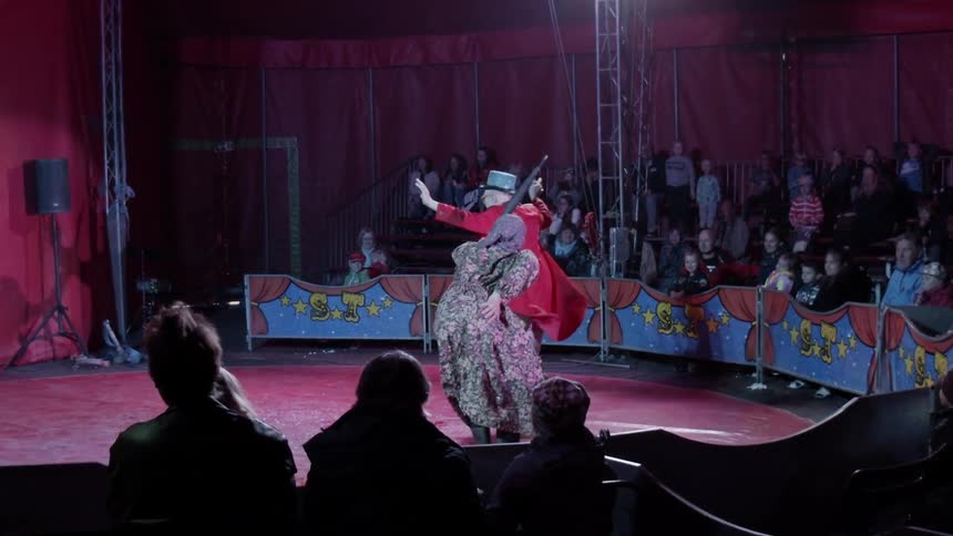 An old lady with a cane chases a man in a red coat with a top hat in front of an audience