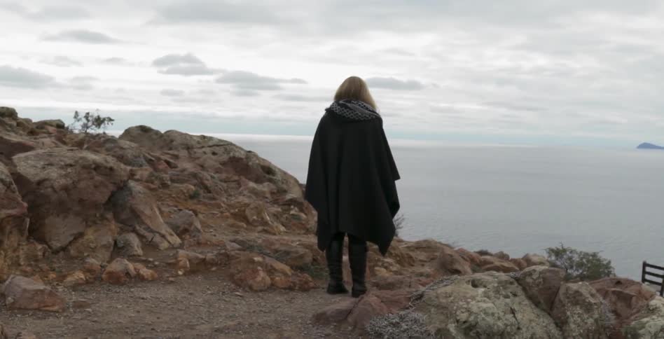A woman stands at a cliffside peak overlooking the ocean