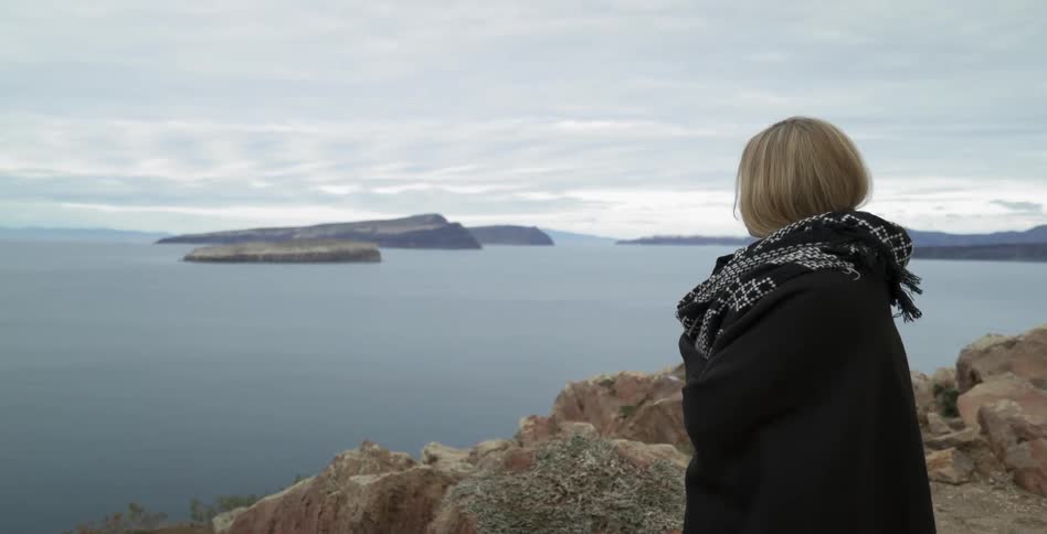 A young woman looks outwards at the shoreline from a cliffside peak