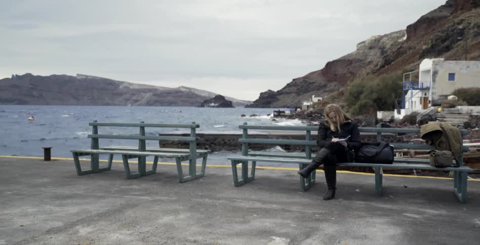A woman sits on a bench by the water writing