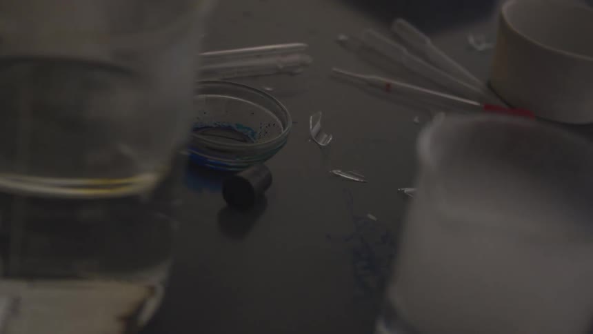 A dark work surface is littered with broken pieces of glass and pipettes while some beakers of smoldering chemicals are nearby