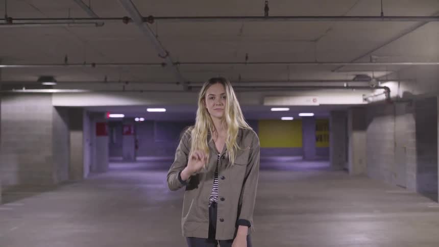 A blonde woman in a car park looks up to the camera and smiles