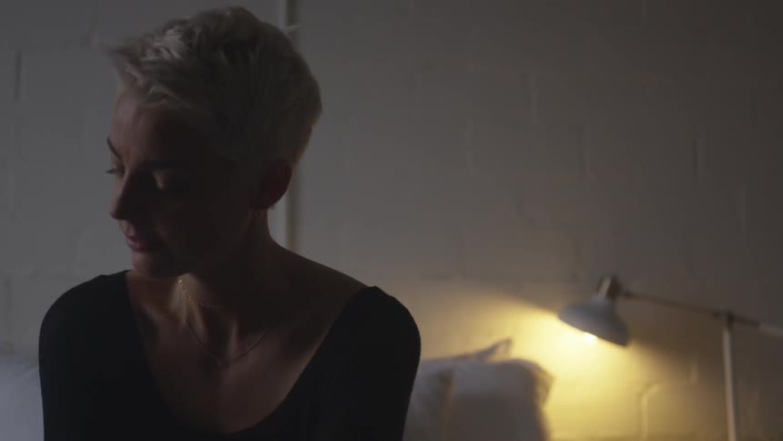 A woman with short hair sits on the bed and closes her eyes while she turns her head