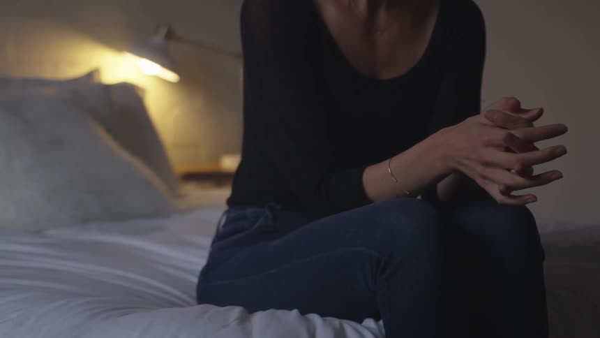 A woman sits on the edge of a bed with her hands clasped together