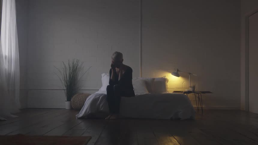 A woman sits on a bed and puts her head in her hands