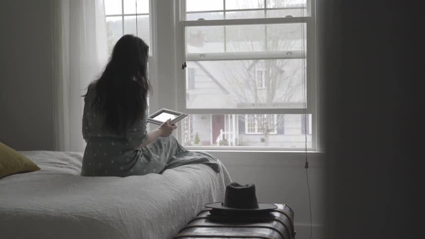 Woman looks at a picture while sitting on a bed before a window
