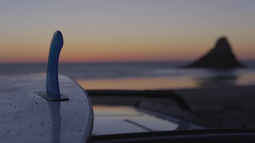 A surfboard over the roof of a car at sunset next to a beach