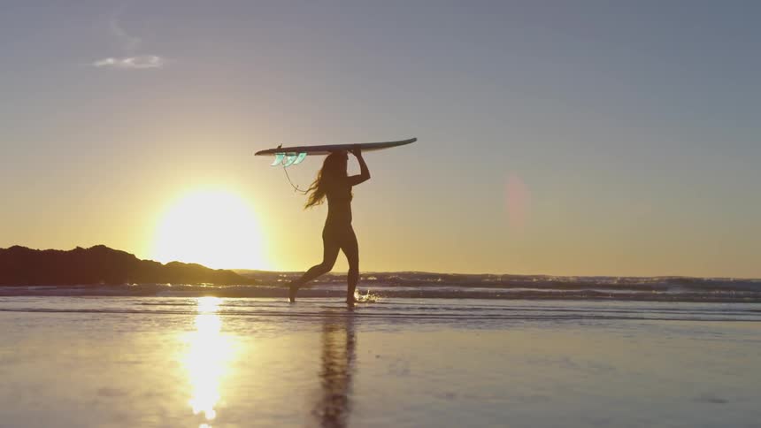 A long haired woman in a bikini is walking across a beach at sunset with a surfboard on her head