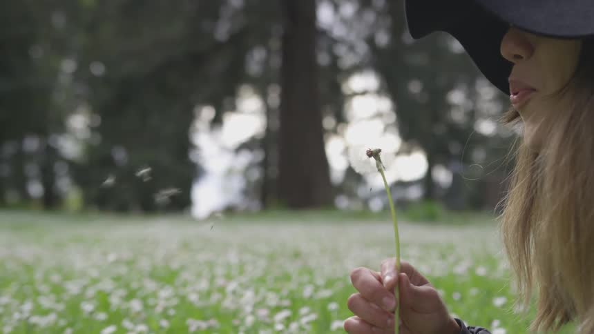 A woman in a hat blowing the petals off of a dandelion