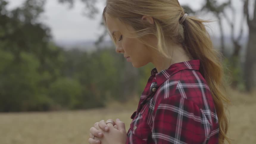 Blonde woman who wears red striped shirt clasps hands and prays in the forest by day