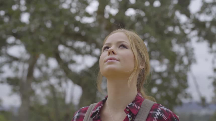 Women stands in a forest looks around upwards up at the trees while wearing a backpack and a necklace