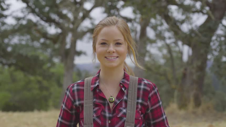 A young girl in overalls and a flannel shirt is looking down and then at the camera