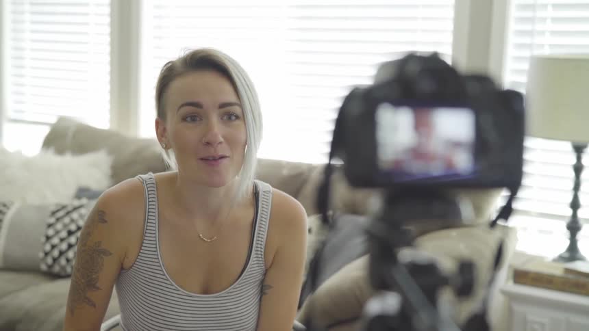 A woman sits and talks in front of a camera on a tripod in a living room