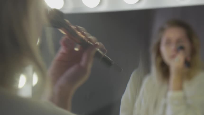 A woman uses a brush to apply makeup to her face while looking at her reflection in a lit mirror