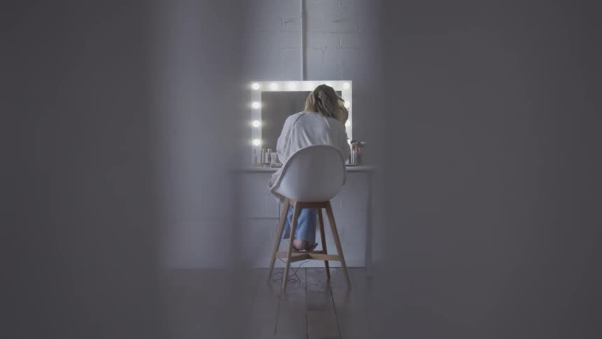 A woman sits in front of a lit up mirror fixing her hair