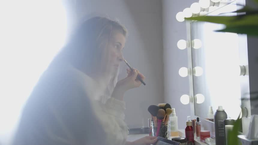 A woman applies makeup under her eye using a brush and then sits back and looks at herself in the mirror