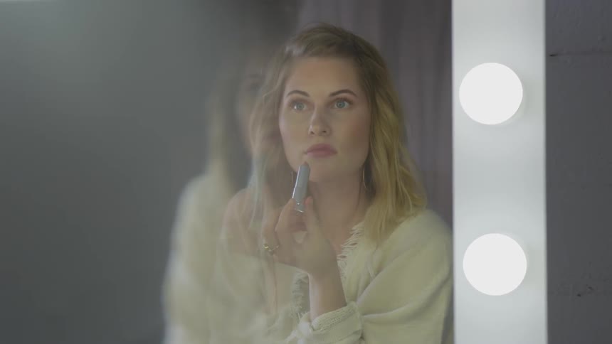 Blonde woman looks in the mirror and puts on lipstick indoors