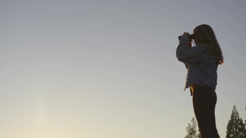 Woman uses binoculars to look towards the sunset