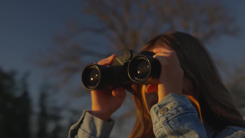 A woman wearing a denim jacket looks out into the sunset using binoculars