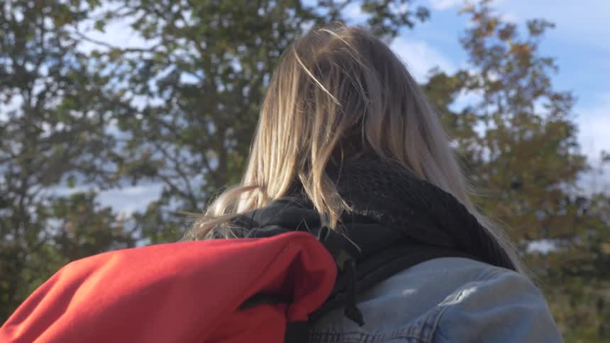 Blonde girl wearing a jacket and a backpack walking nearby trees