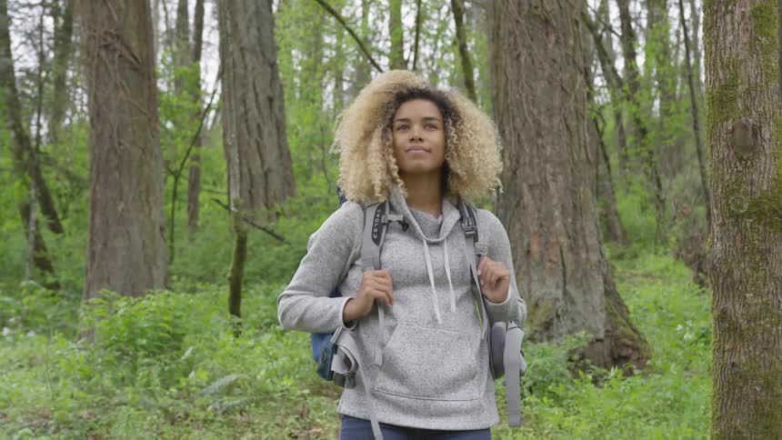 She's got blonde dyed hair like a surfer she has a backpack and she is standing in the forest alone looking around with a surreal look on her face
