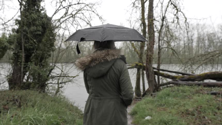 A woman wearing a parka and holding an umbrella stands on a grassy river bank looking at the river