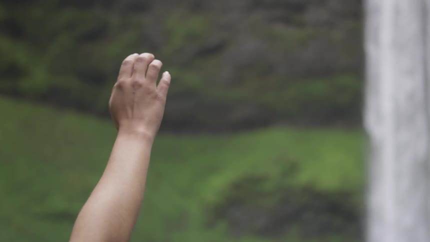 A person is raising their hand outdoors by a waterfall